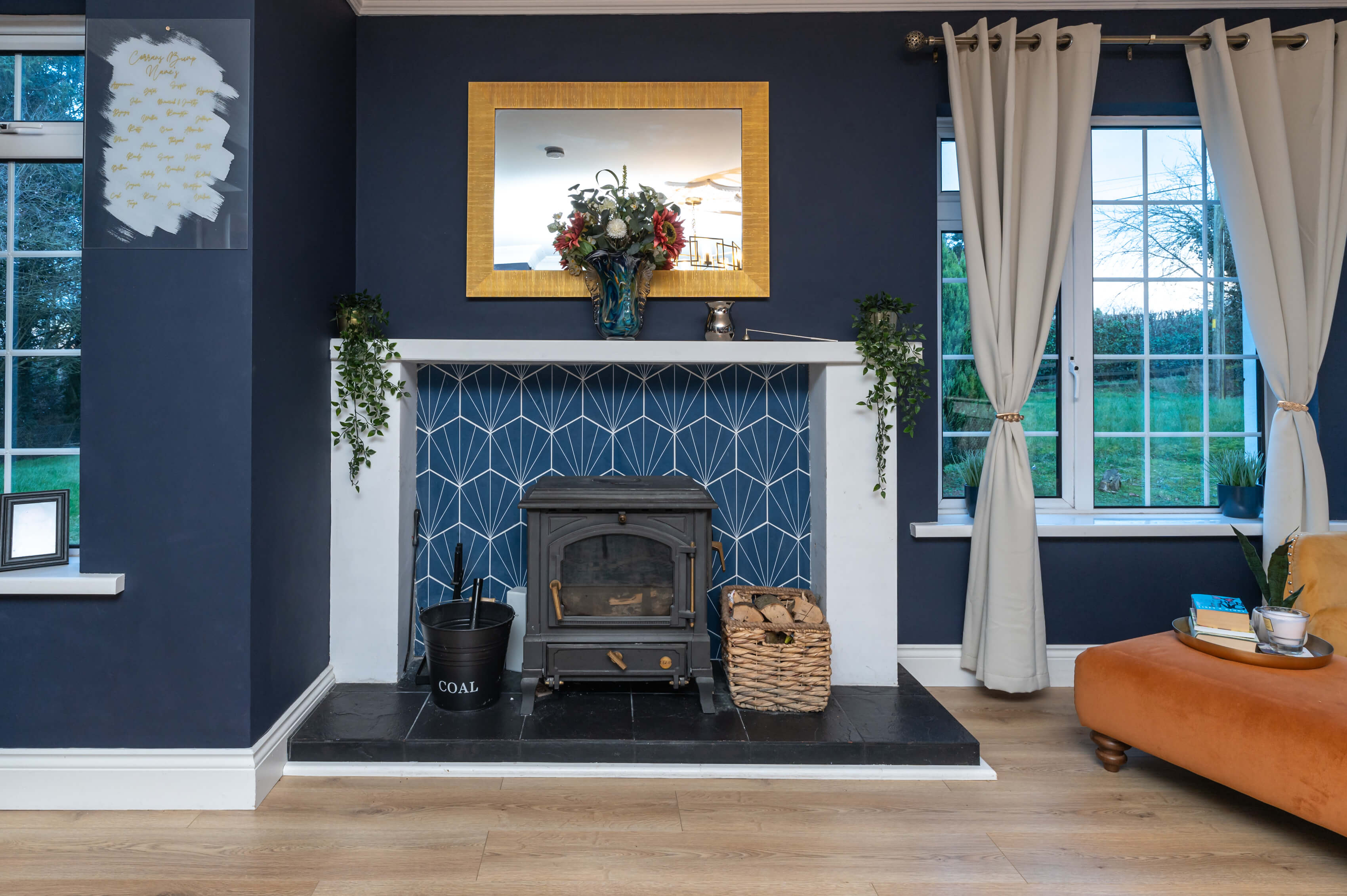 a living room fireplace with white surround, blue geometric tiles and a black stove. There is a gold mirror above the fire place, the walls are dark navy. Faux plants hang either end of the mantlepiece. The curtains are a neutral colour with a brushed gold curtain pole. A deep yellow accent chair and orange footstool are to the side of the fireplace.